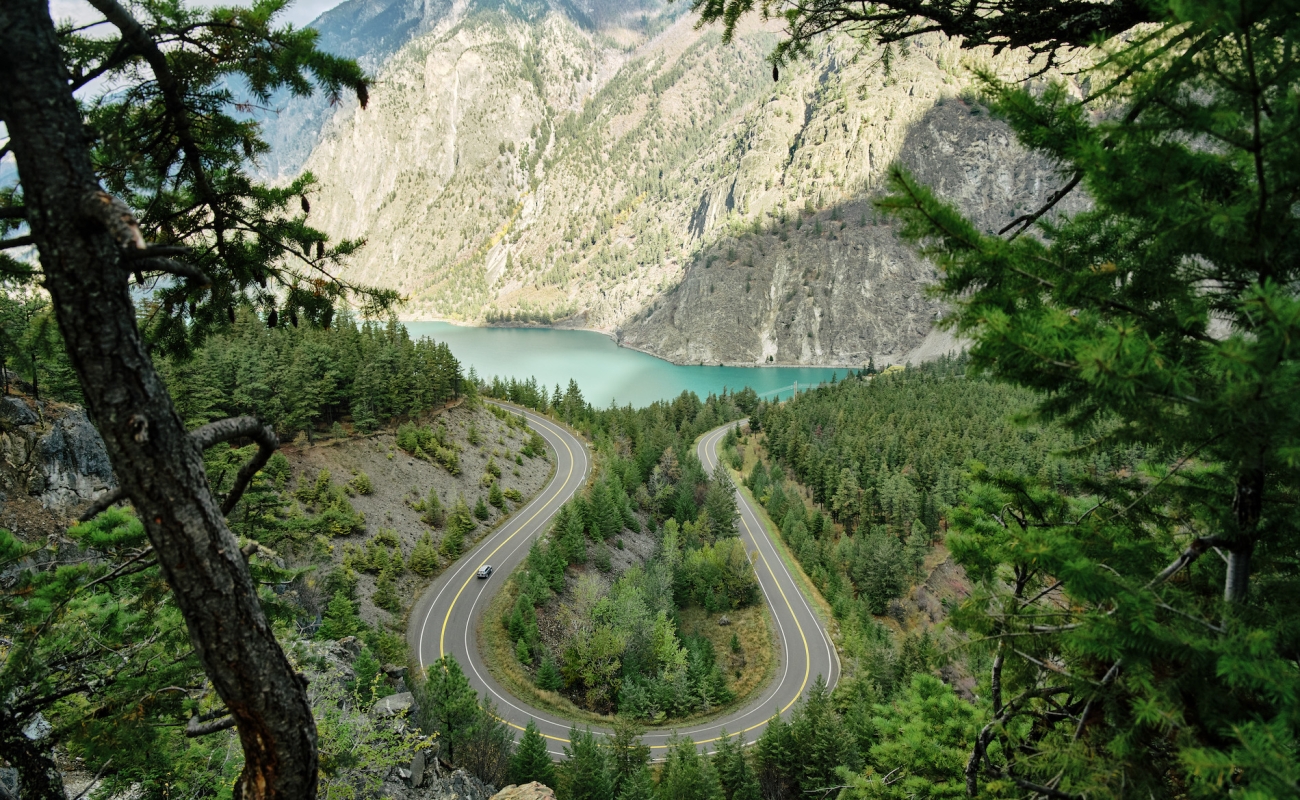 Vista hacia un lago turquesa con un paisaje boscoso en primer plano y montañas cubiertas de artemisa al fondo.