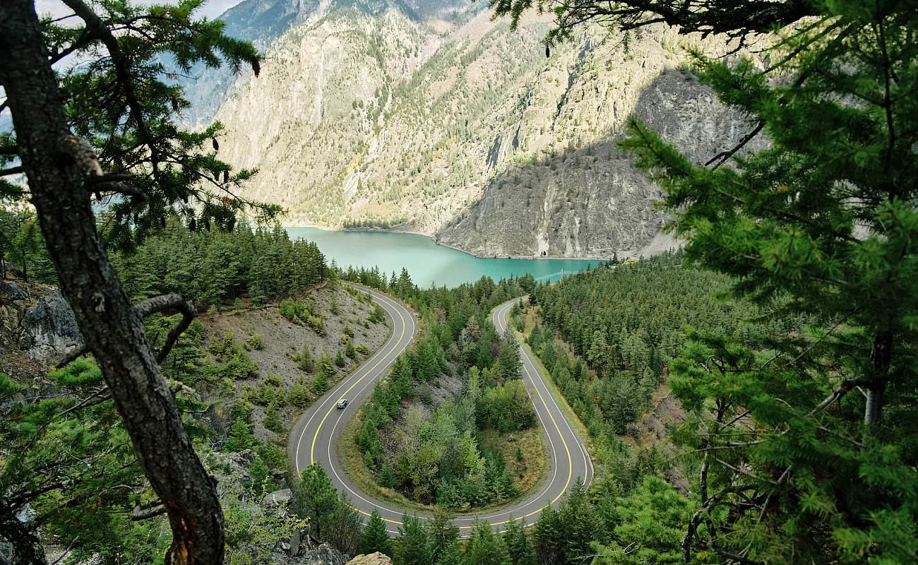 Blick auf einen türkisfarbenen See mit einer bewaldeten Landschaft im Vordergrund und mit Salbeibüschen bewachsenen Bergen im Hintergrund.