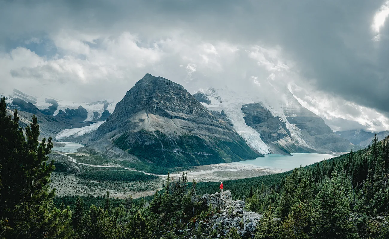 Eine Person sitzt auf einem Felsvorsprung und blickt auf die hoch aufragenden Gipfel und riesigen Gletscher des Mt. Robson.