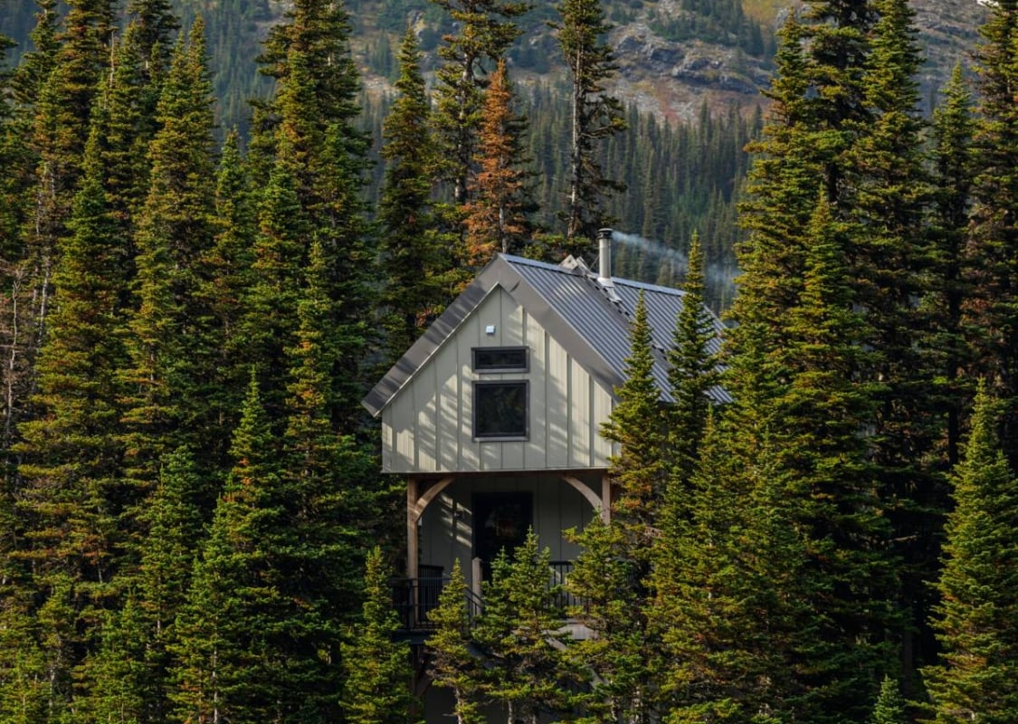 A quaint cabin is tucked in a forest overlooking a lake with towering mountains in the background.