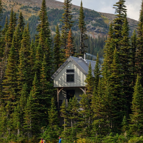 A quaint cabin is tucked in a forest overlooking a lake with towering mountains in the background.