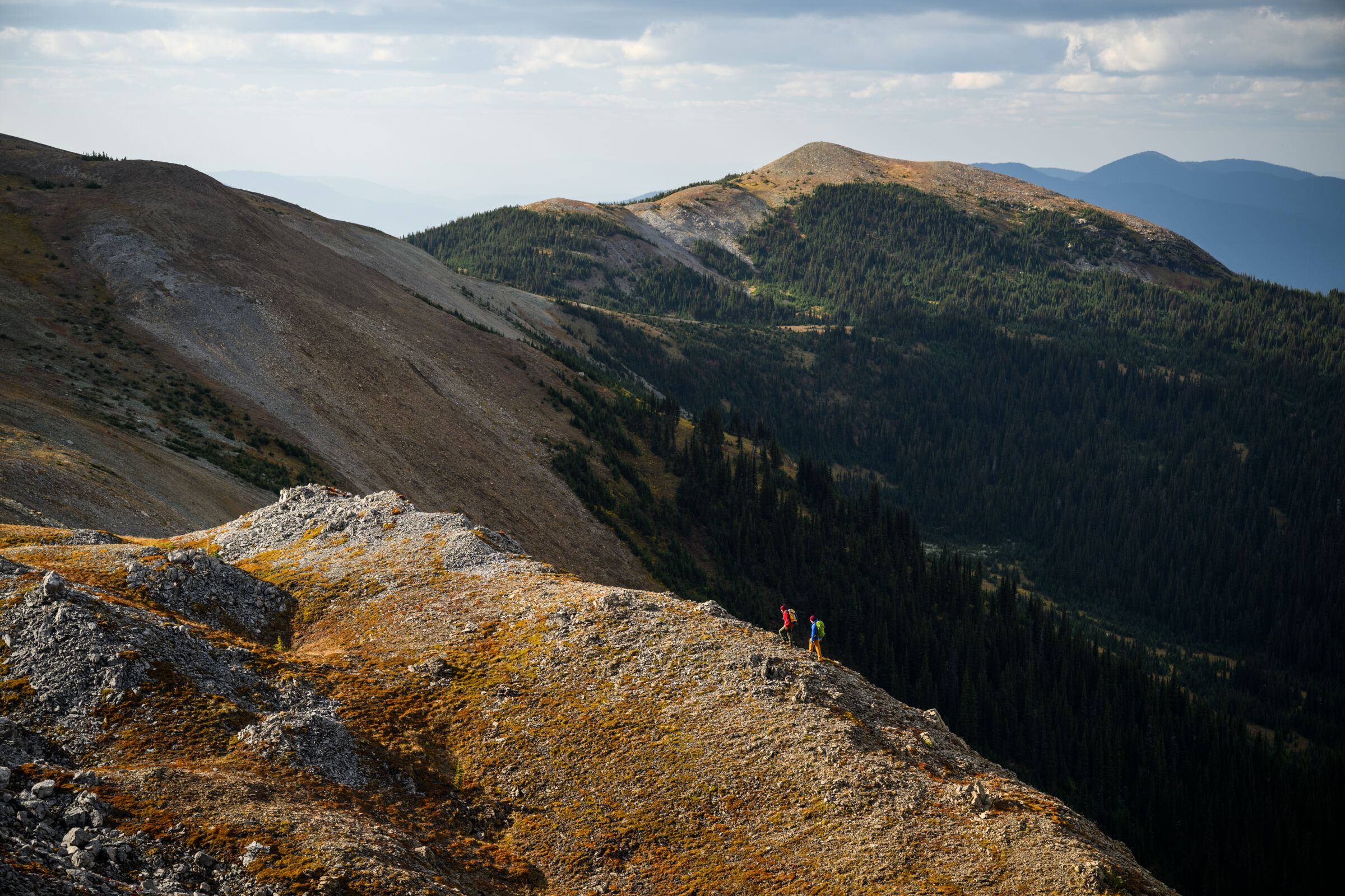 Two hikers walk across a rocky ridge overlooking orange mountains dotted by lush green forest.