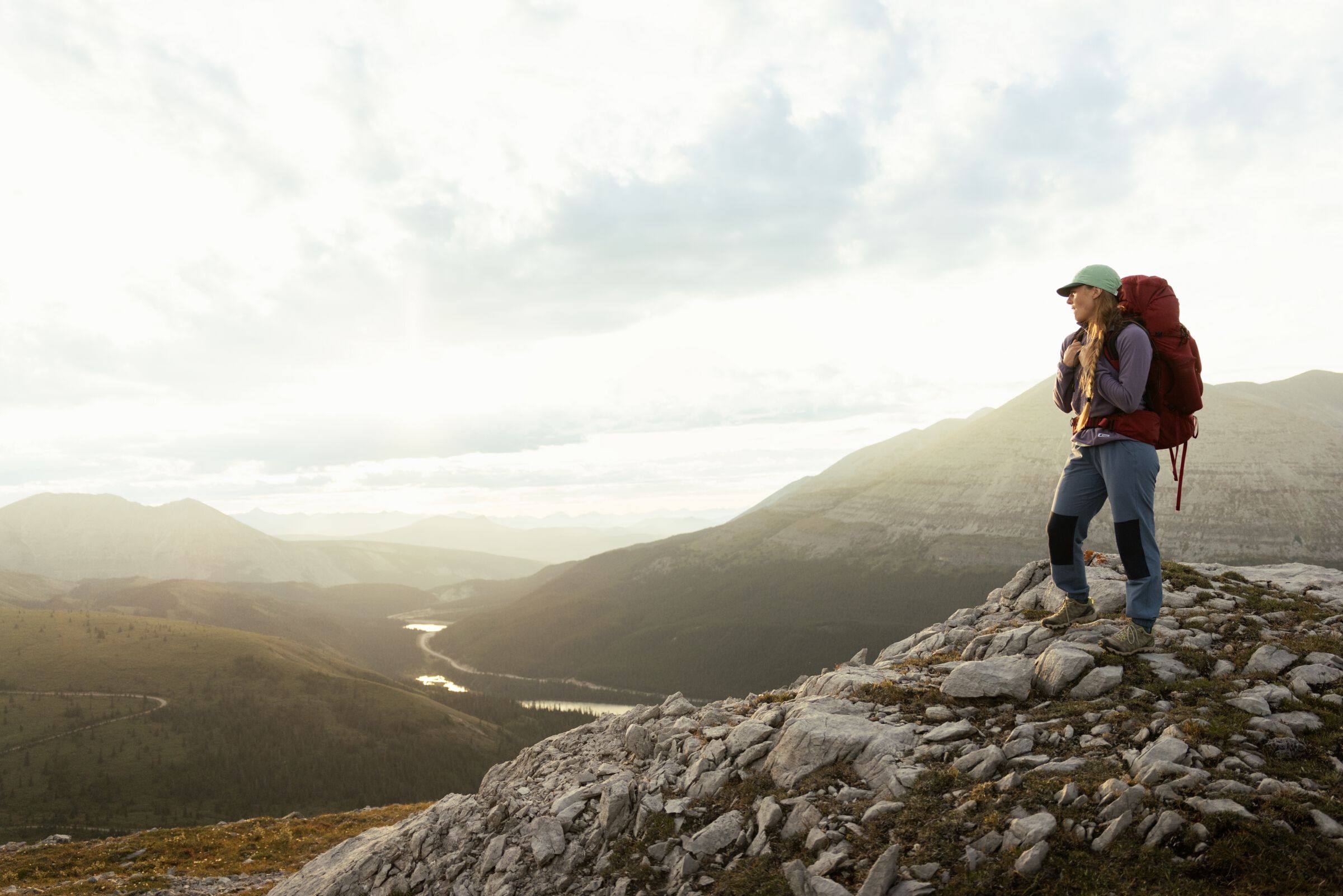 A person wearing hiking gear and a backpack stands on a rocky outcropping overlooking a river valley with mountains in the background.