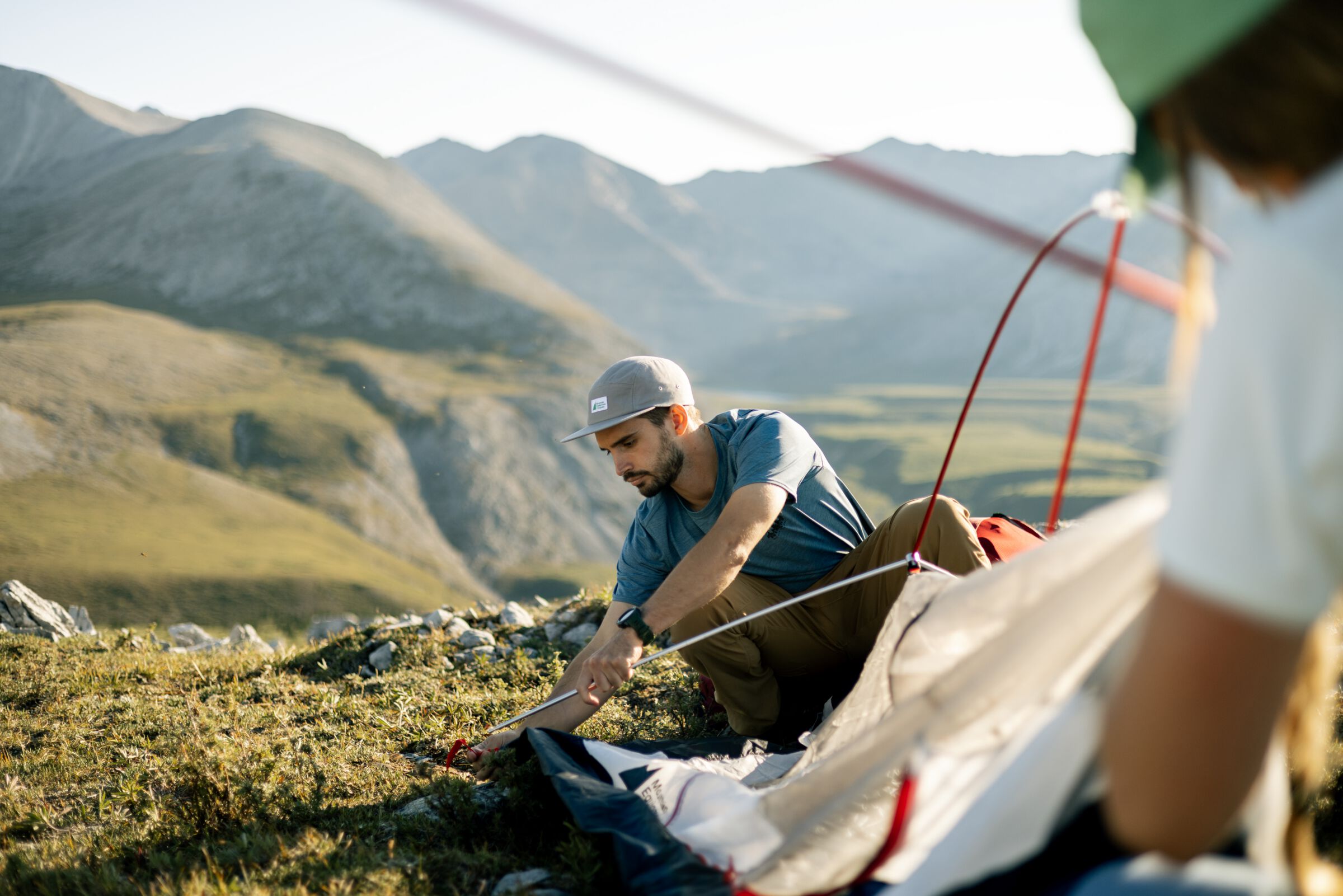 Two people set up a backcountry tent on a grassy landscape with mountains in the background.