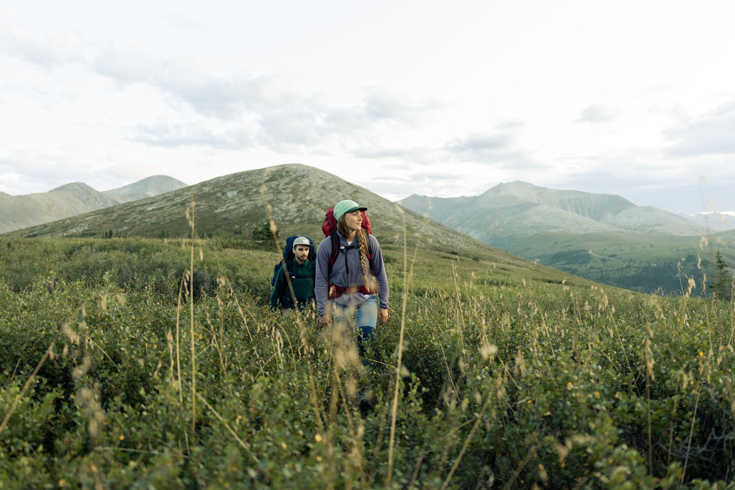 Two people with hiking backpacks trek through a lush meadow with rolling foothills in the background.