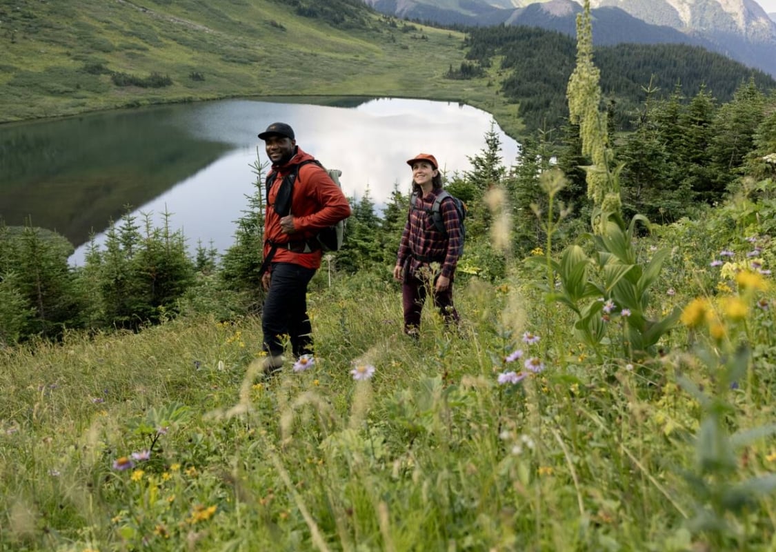 Two people walk beside an alpine lake, located in a lush meadow of green foliage with craggy mountain tops peeking out behind.