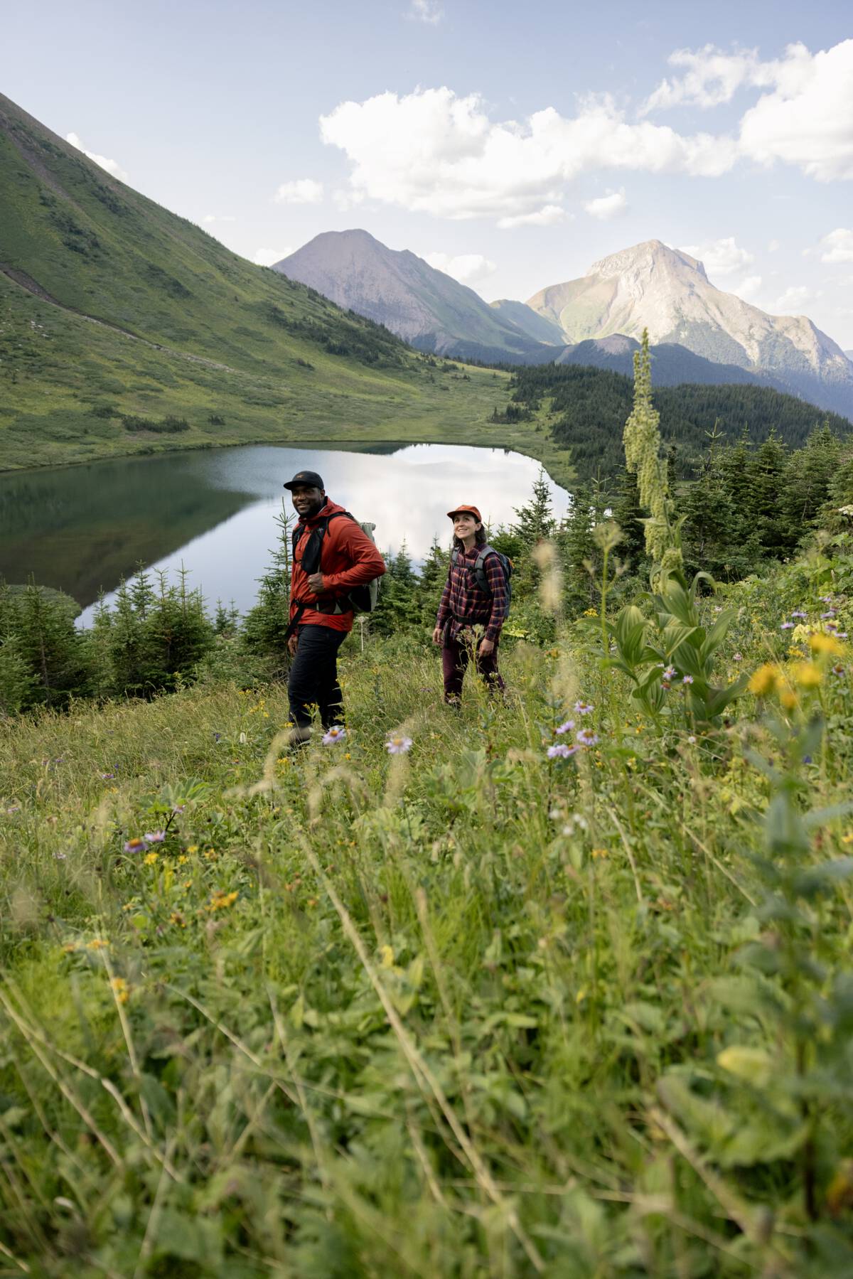 Two people walk beside an alpine lake, located in a lush meadow of green foliage with craggy mountain tops peeking out behind.