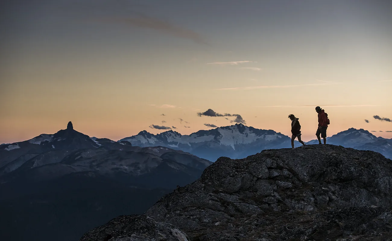 Zwei Wander*innen stehen bei Sonnenuntergang auf dem High Note Trail. In der Ferne ist Black Tusk zu sehen.