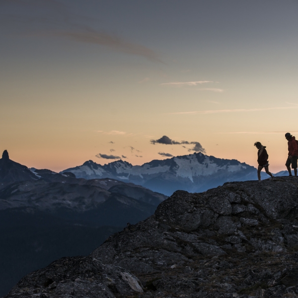 Zwei Wander*innen stehen bei Sonnenuntergang auf dem High Note Trail. In der Ferne ist Black Tusk zu sehen.
