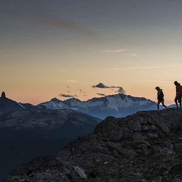 Zwei Wander*innen stehen bei Sonnenuntergang auf dem High Note Trail. In der Ferne ist Black Tusk zu sehen.