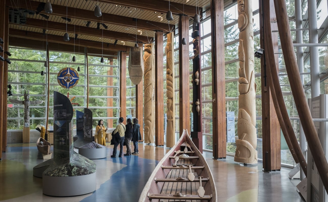 Un grupo de visitantes explora el Squamish Lil'wat Cultural Centre con un guía turístico. Están de pie en un gran salón con ventanas a ambos lados y una canoa de madera en el centro.