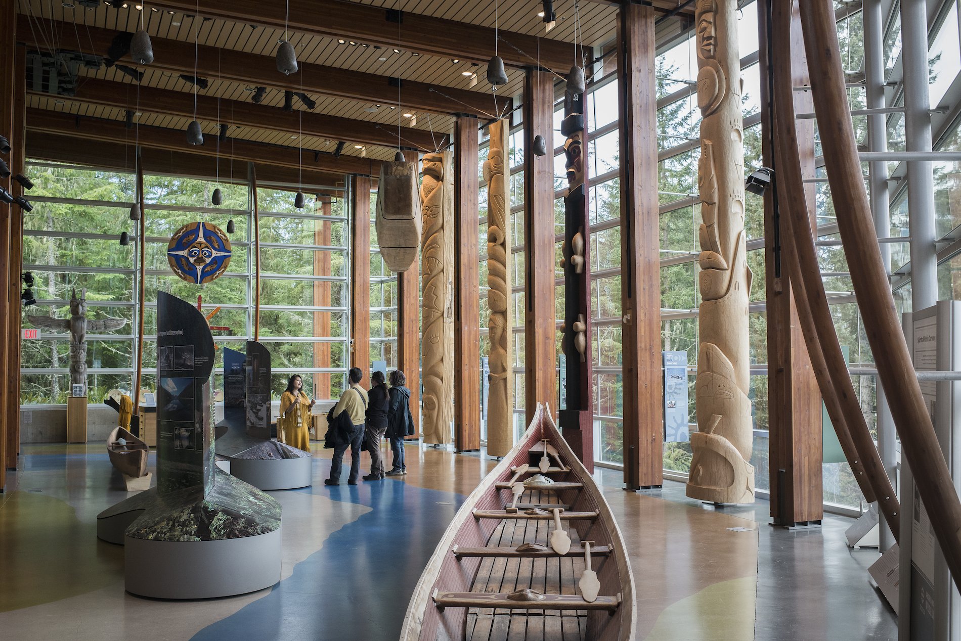 A group of visitors explore the Squamish Lil'wat Cultural Centre with a tour guide. They stand in a large hall with windows on both sides and a wooden canoe in the centre.