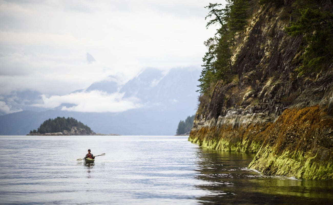 A person kayaks around the Sunshine Coast. A rocky shoreline is next to the paddler in the distance.