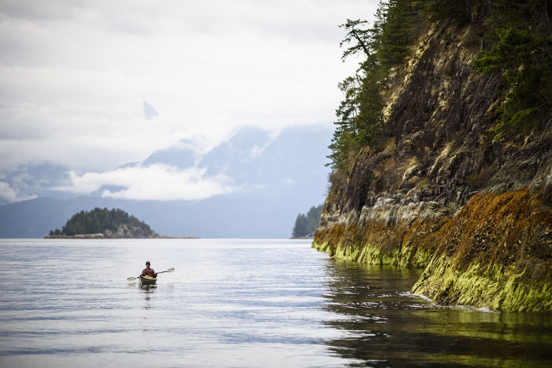 A person kayaks around the Sunshine Coast. A rocky shoreline is next to the paddler in the distance.