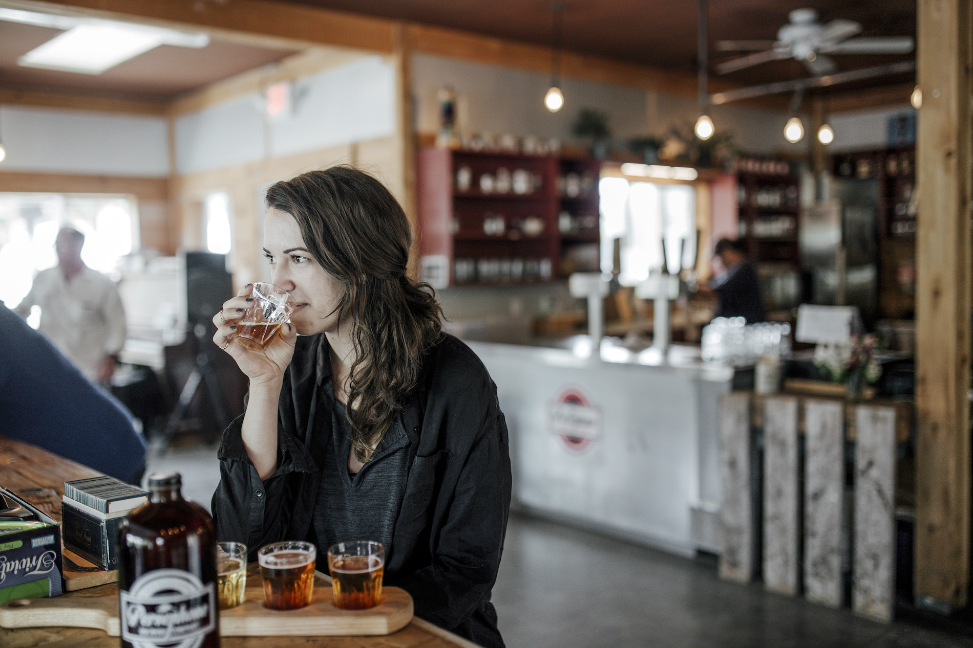 A woman sips on a beer at the Persephone Brewing Company. There is a flight of beers in front of her on the table. Behind her is the counter with beers on tap.