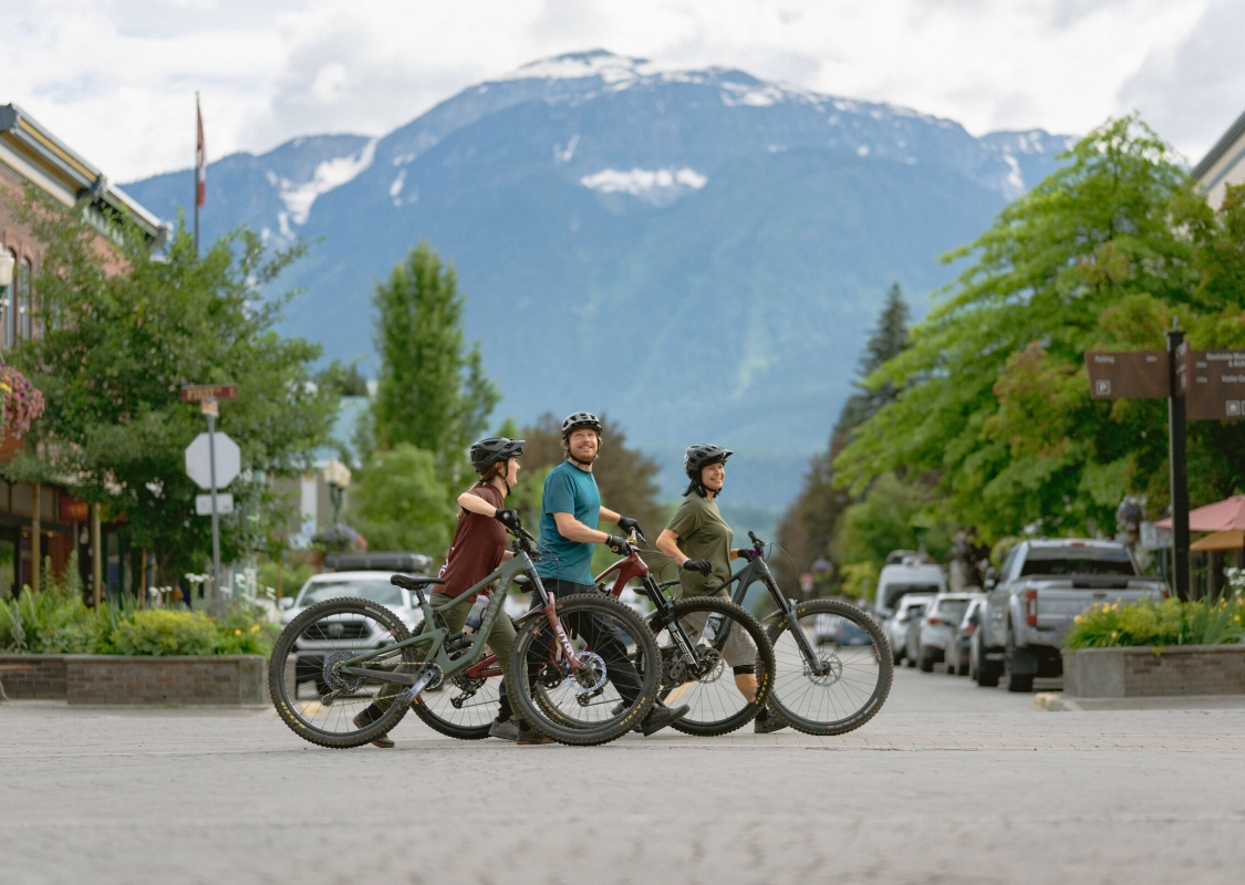 Three friends walking their bikes across the street during their bike ride in downtown Revelstoke.
