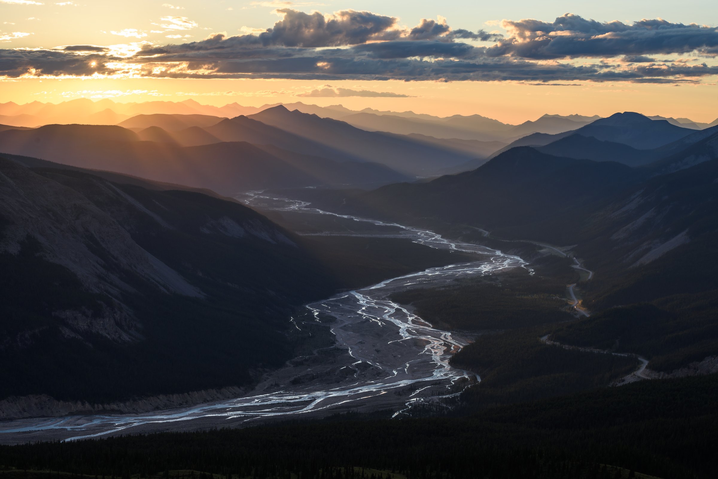Sunset over a mountain valley with a river slicing through the centre as rays of light dance across the peaks.