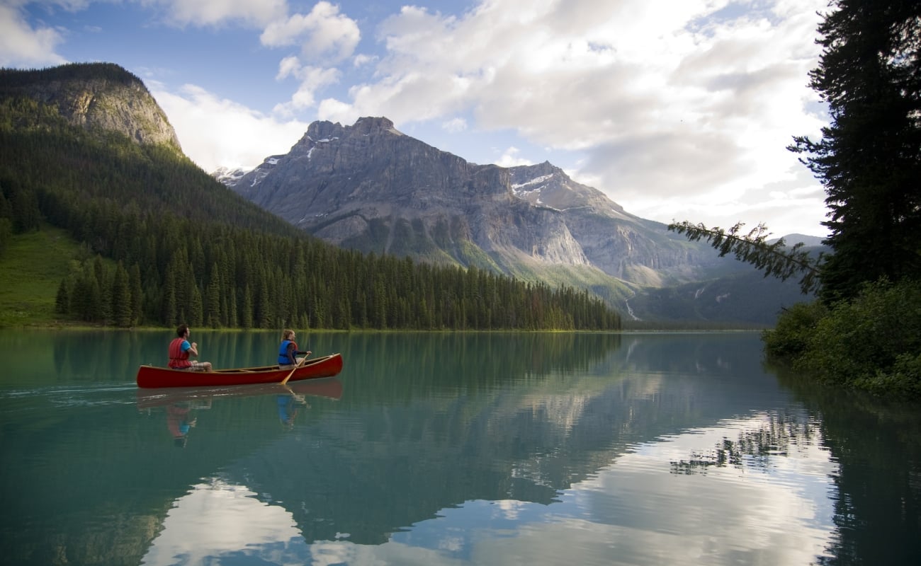 Ein rotes Kanu wird von zwei Personen über einen See im Yoho National Park gepaddelt. Der türkisfarbene See ist von Bergen und Bäumen umgeben.