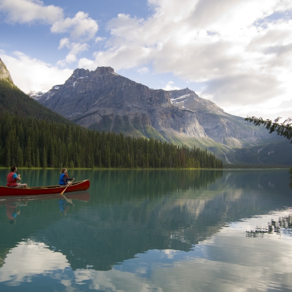 A red canoe is paddled by two people across a lake in Yoho National Park. The turquoise lake is surrounded by mountains and trees.