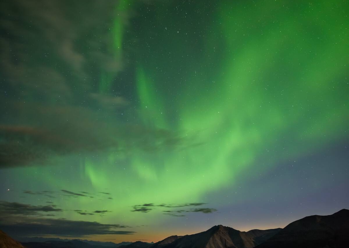 A yellow tent lit up by interior lights shines underneath a starry sky as vibrant green Aurora Borealis dance across the sky.