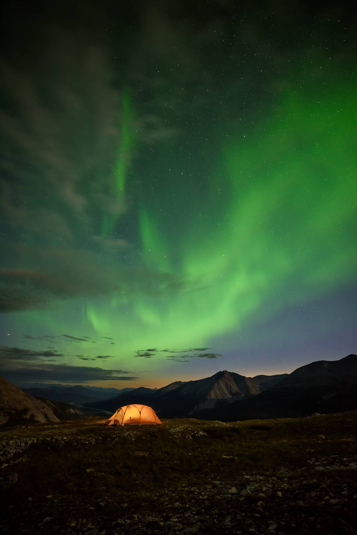 A yellow tent lit up by interior lights shines underneath a starry sky as vibrant green Aurora Borealis dance across the sky.