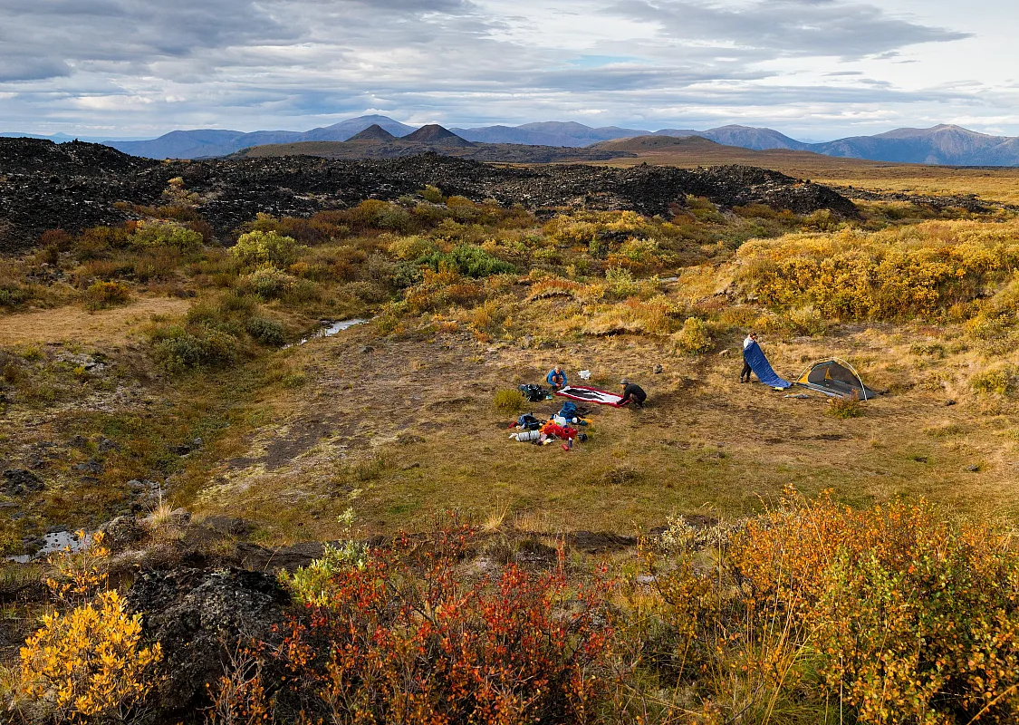 A group sets up their tents in an alpine meadow with various shades of green, yellow and orange foliage and mountains in the background.