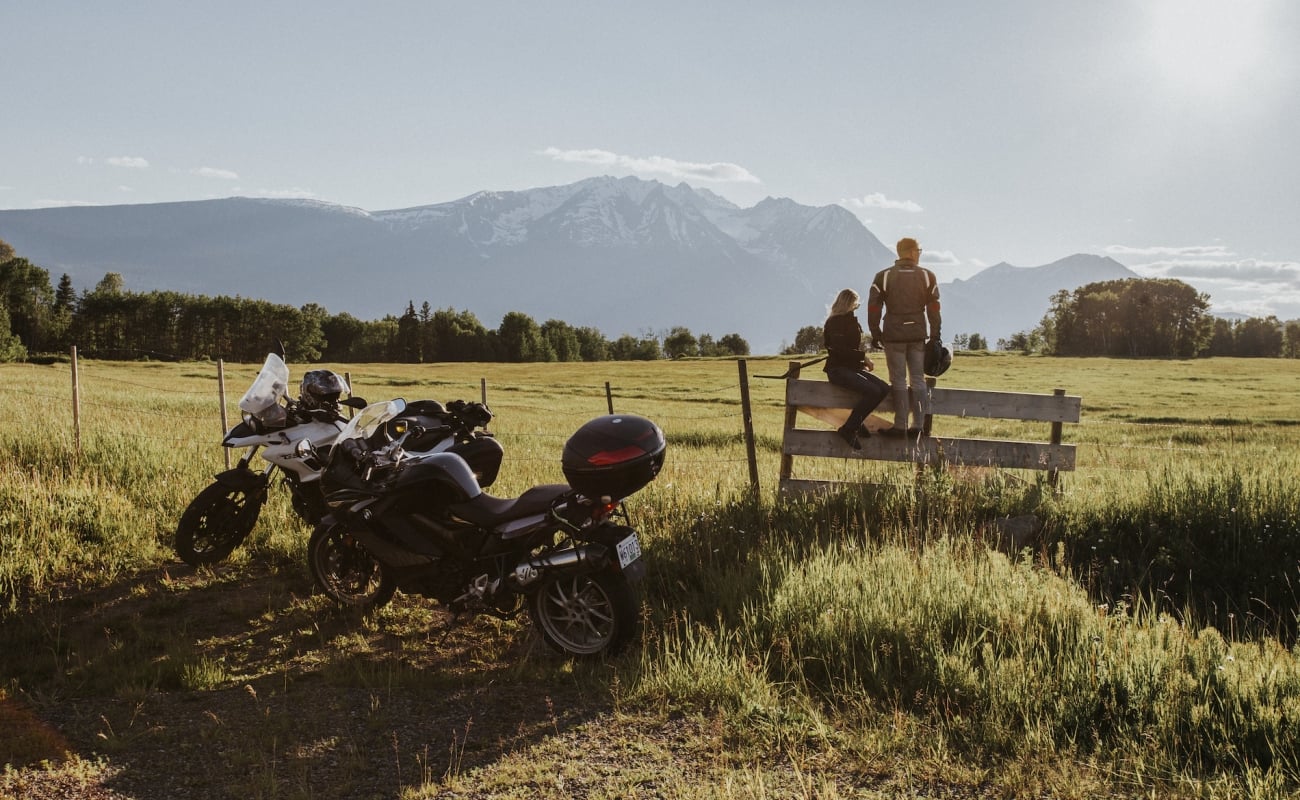 Zwei Motorräder stehen vor einem Feld, die Fahrer*innen sitzen auf einem Zaun.