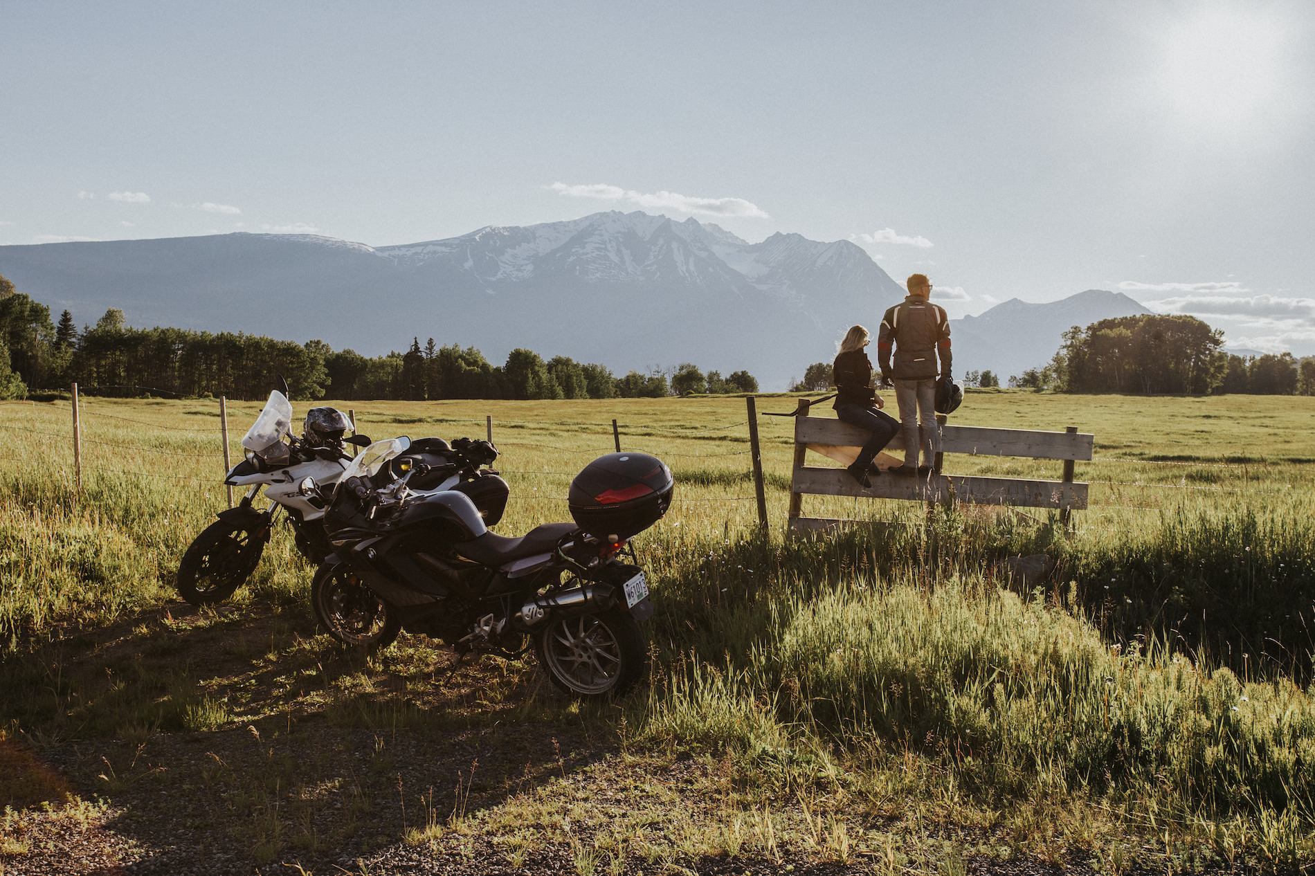Two motorcycles are parked in front of a farm field with the riders sitting on a fence.