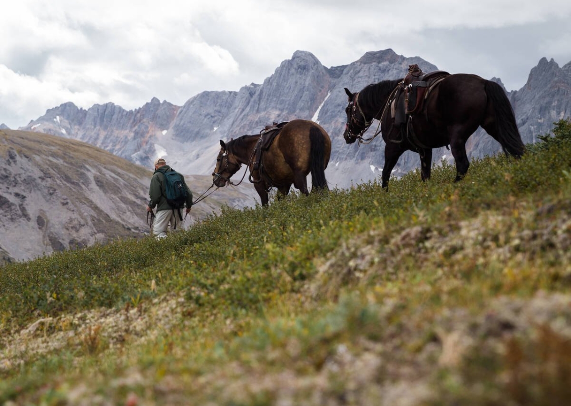 A horseback guide leads two horses across a grassy foothill with rugged mountains in the background.
