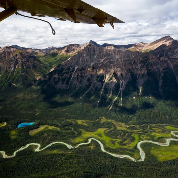 Ein Flugzeugflügel ragt über eine Luftaufnahme eines leuchtend grünen Tals, durch dessen Mitte sich ein Fluss schlängelt, umgeben von steil abfallenden Bergen.