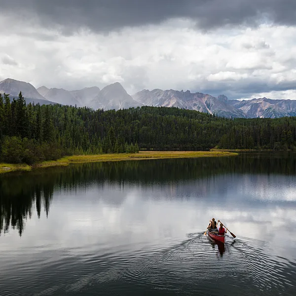 Two people paddle a red canoe across a lake toward a forest with mountains in the backgorund.