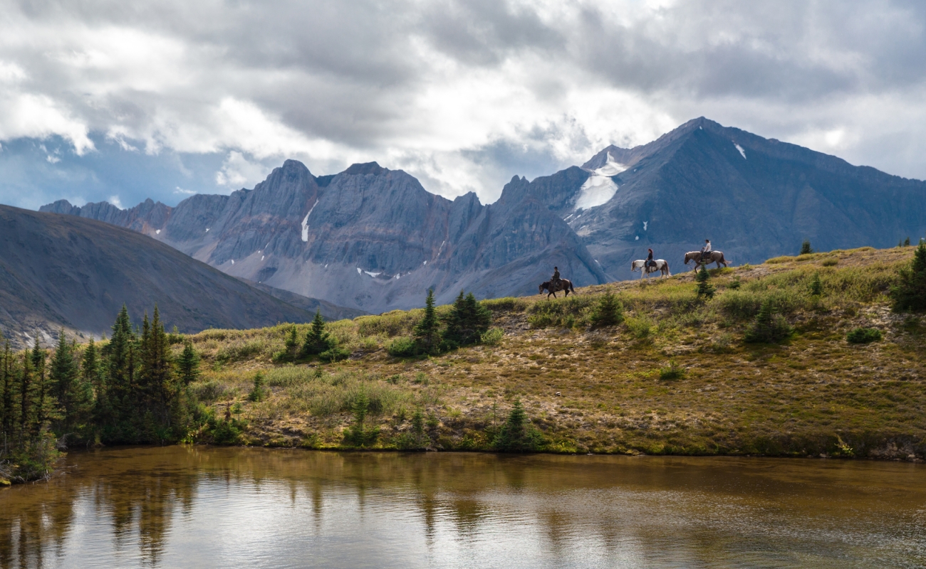 A group of horseback riders roams the vast countryside with mountains in the background and river in the foreground.