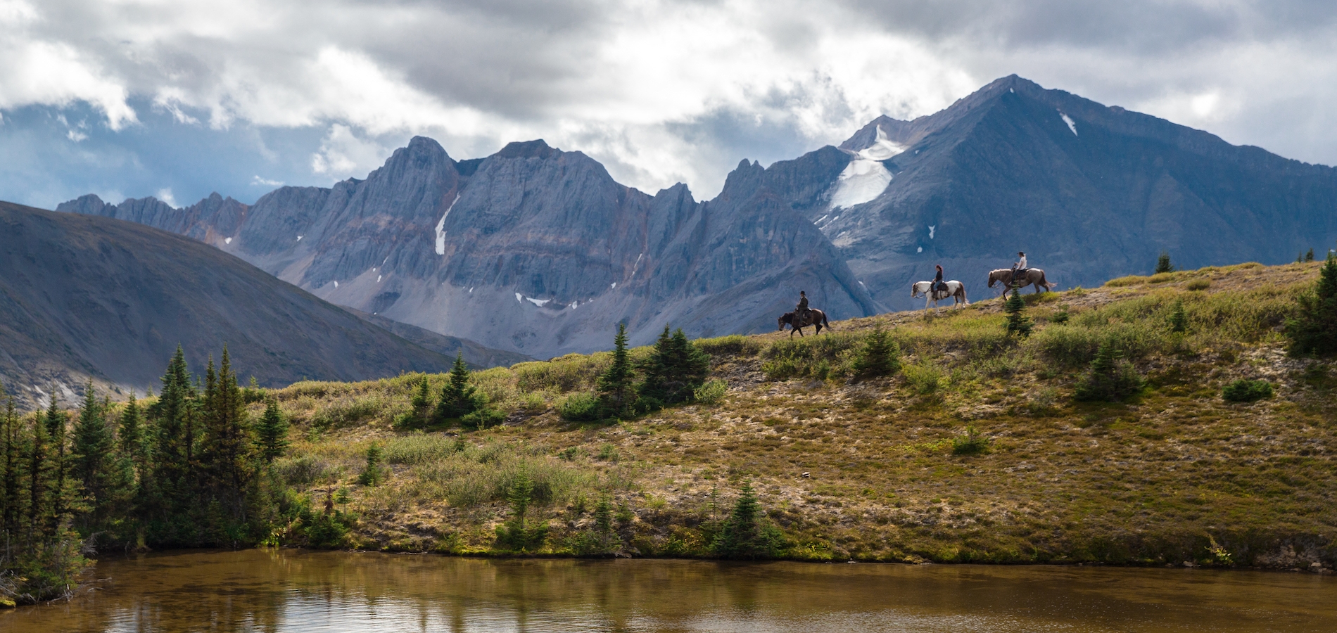 A group of horseback riders roams the vast countryside with mountains in the background and river in the foreground.