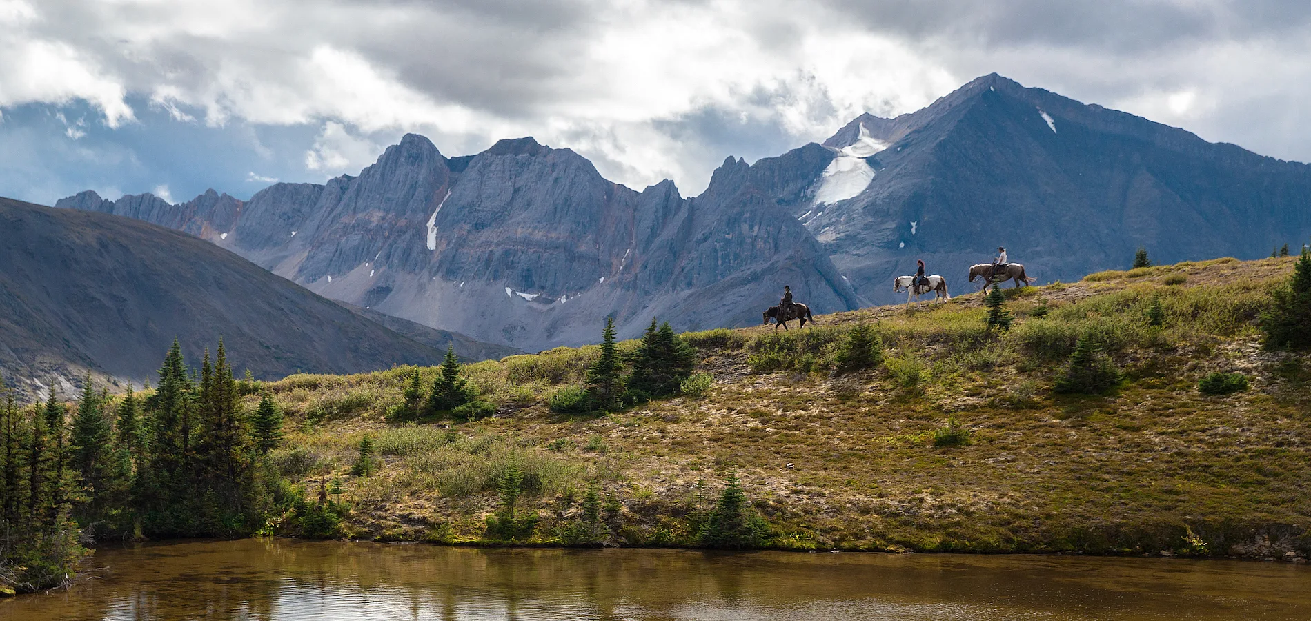 Un grupo de jinetes recorre la vasta campiña con montañas al fondo y un río en primer plano.