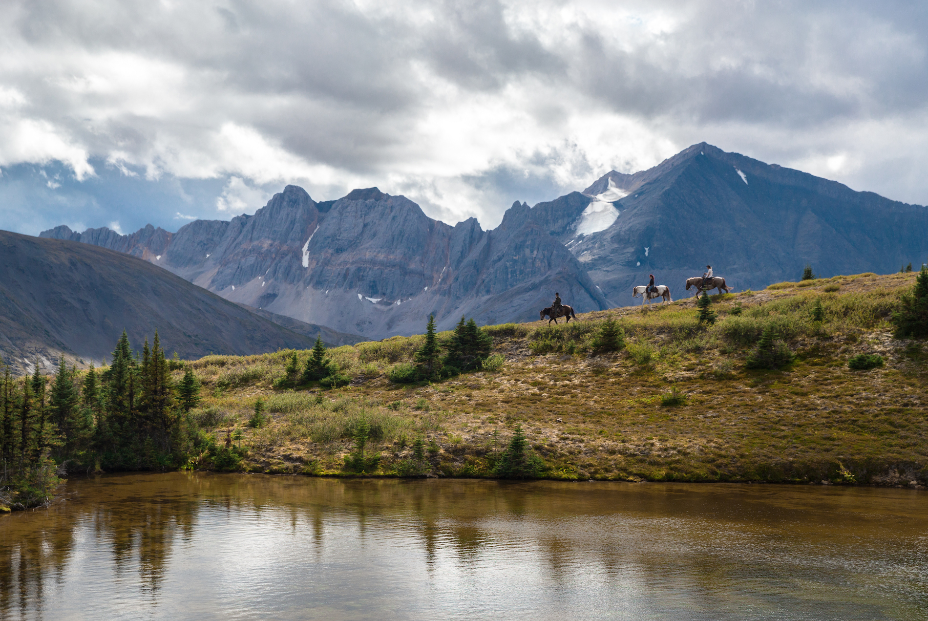 A group of horseback riders roams the vast countryside with mountains in the background and river in the foreground.