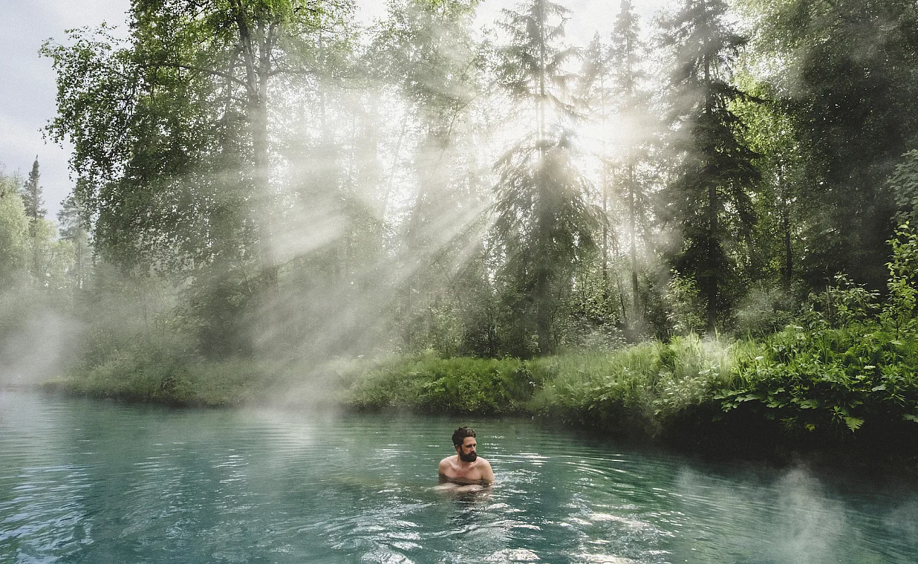 Una persona se relaja en las Liard hot springs con rayos de luz asomándose entre los árboles.