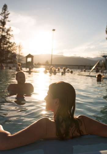 People enjoy a dip in then Fairmont Hot Springs outdoor hot pools. The sun is setting and reflecting across the water. A women in the foreground relaxes by the pool edge with her back to the camera.