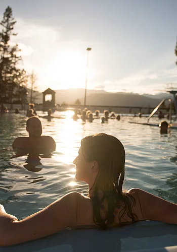 Leute genießen ein Bad in den Fairmont Hot Springs Außenpools. Die Sonne geht unter und spiegelt sich auf dem Wasser. Eine Frau im Vordergrund entspannt sich am Poolrand mit dem Rücken zur Kamera.