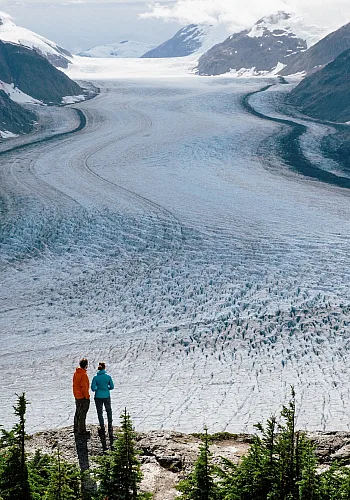 Two people look out at a frozen landscape in the Stewart-Cassiar region of northern BC. They stand on a rocky outcrop and are looking towards the mountain range.