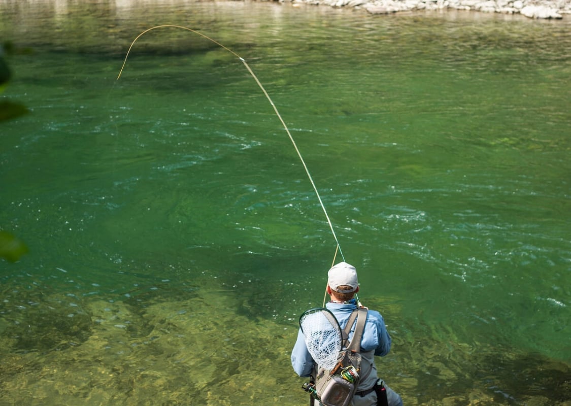 A person stands at a glistening green river with a fly fishing rod.