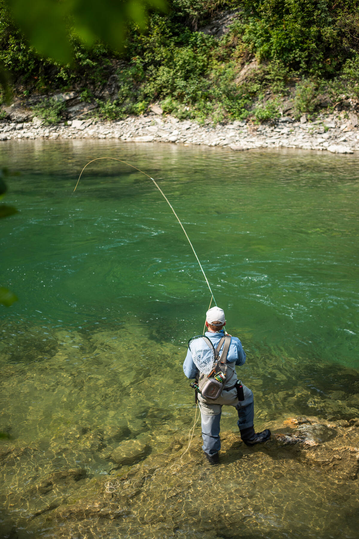 A person stands at a glistening green river with a fly fishing rod.