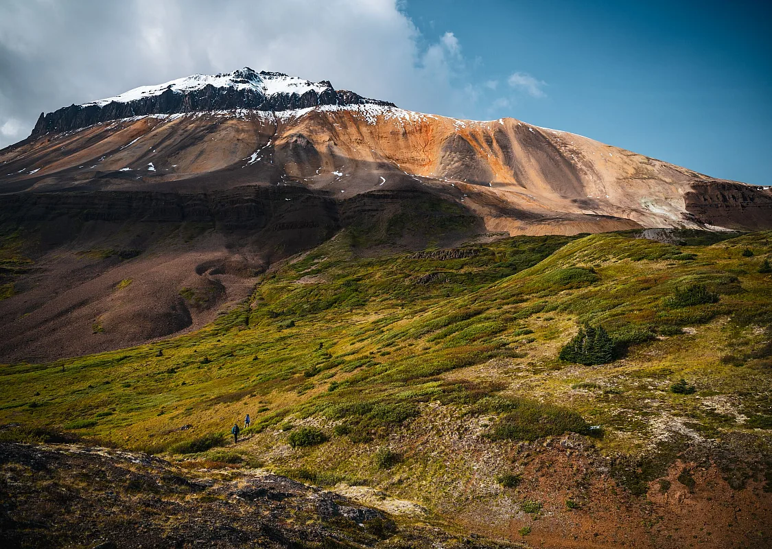 A dramatic and colourful landscape with an orange, mineral-streamed mountain that is covered in snow at the very top, overlooking a lush green alpine meadow.