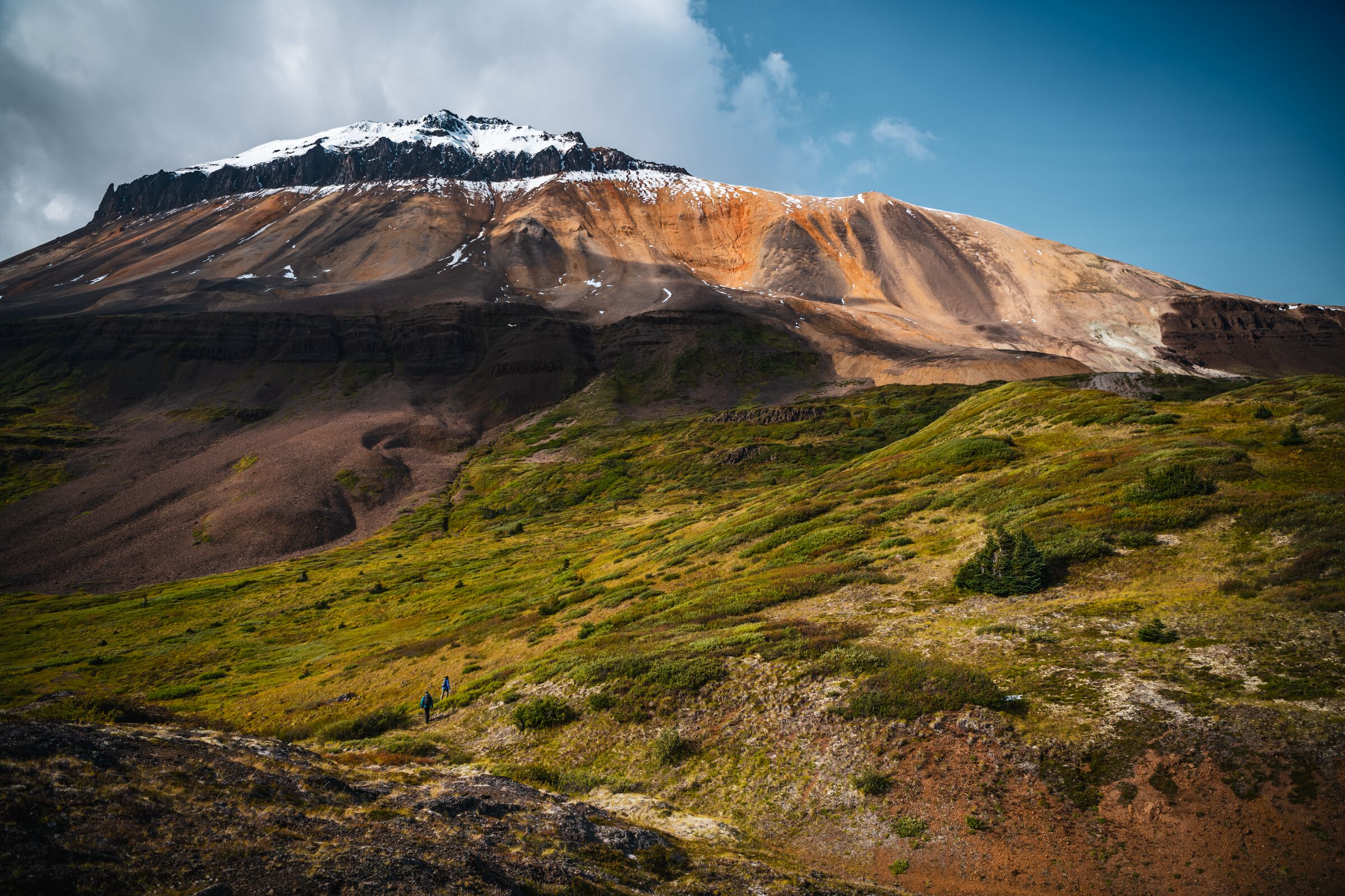 A dramatic and colourful landscape with an orange, mineral-streamed mountain that is covered in snow at the very top, overlooking a lush green alpine meadow.