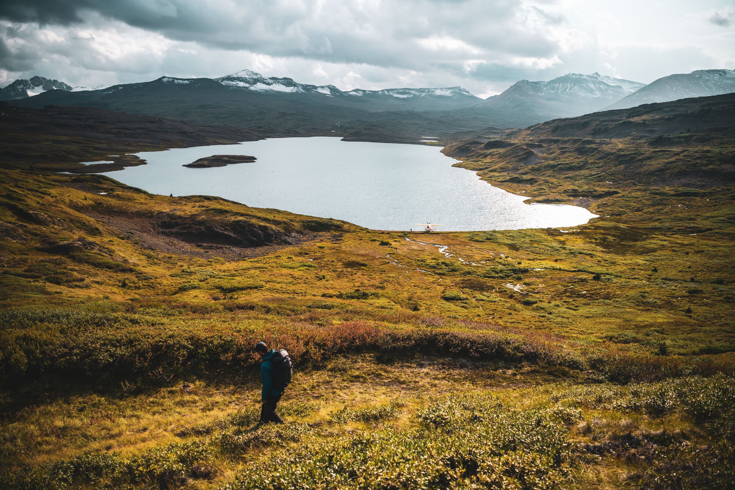 A hiker walks through an alpine meadow with low-lying foliage, mountains in the background, and a small lake in the distance.