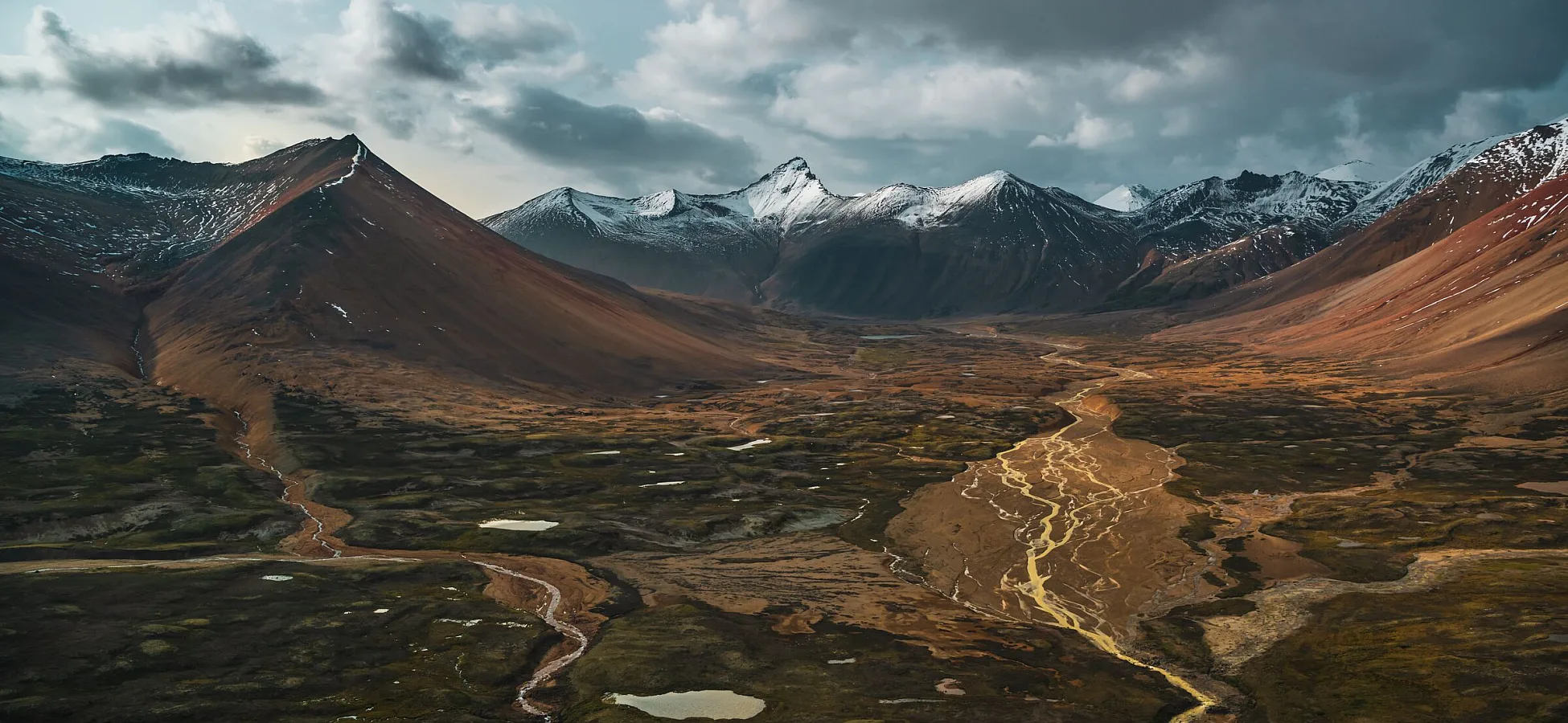 An aerial view of a vast landscape with mineral-streaked orange mountains and a lush river valley below.