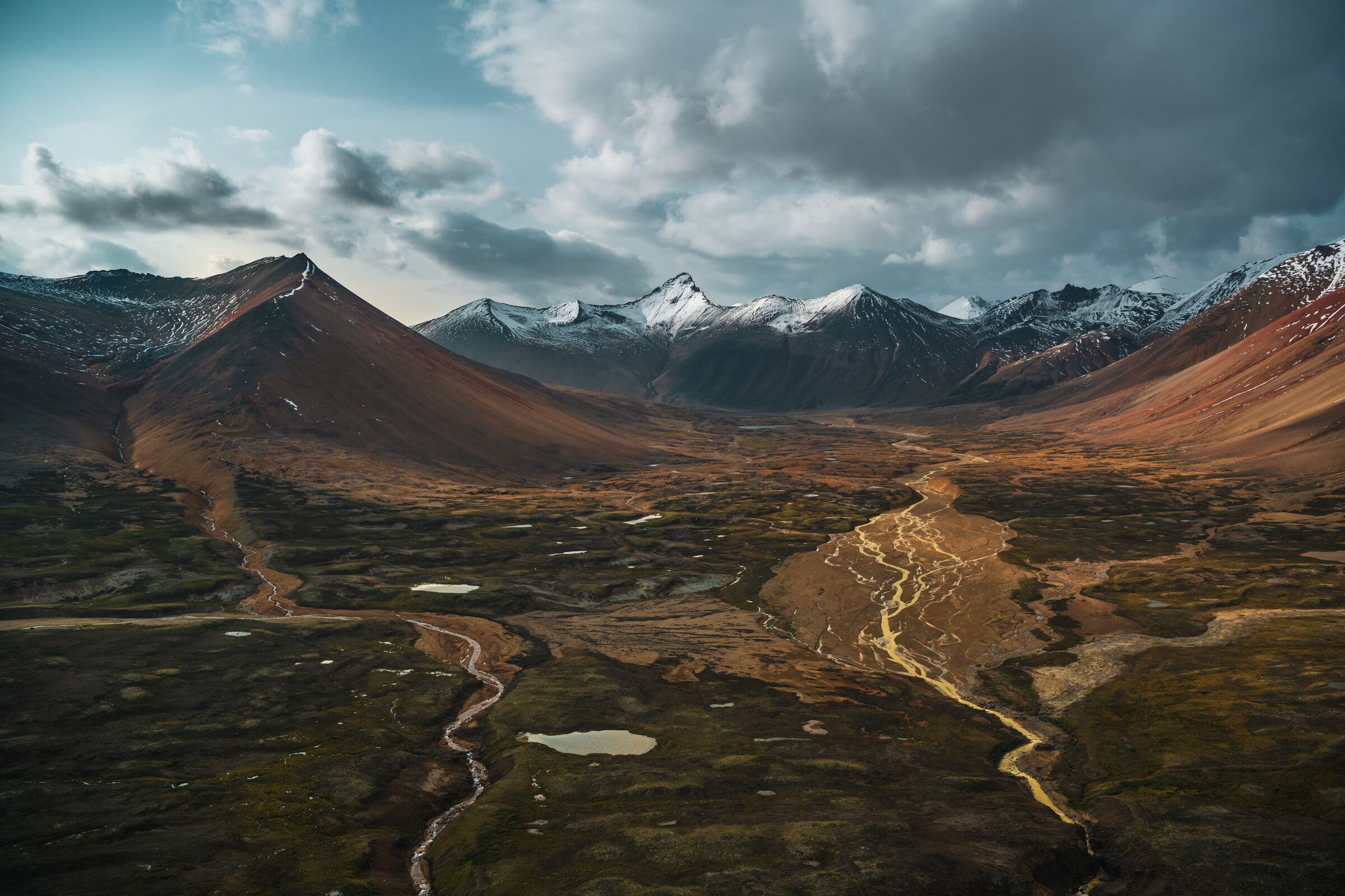 An aerial view of a vast landscape with mineral-streaked orange mountains and a lush river valley below.