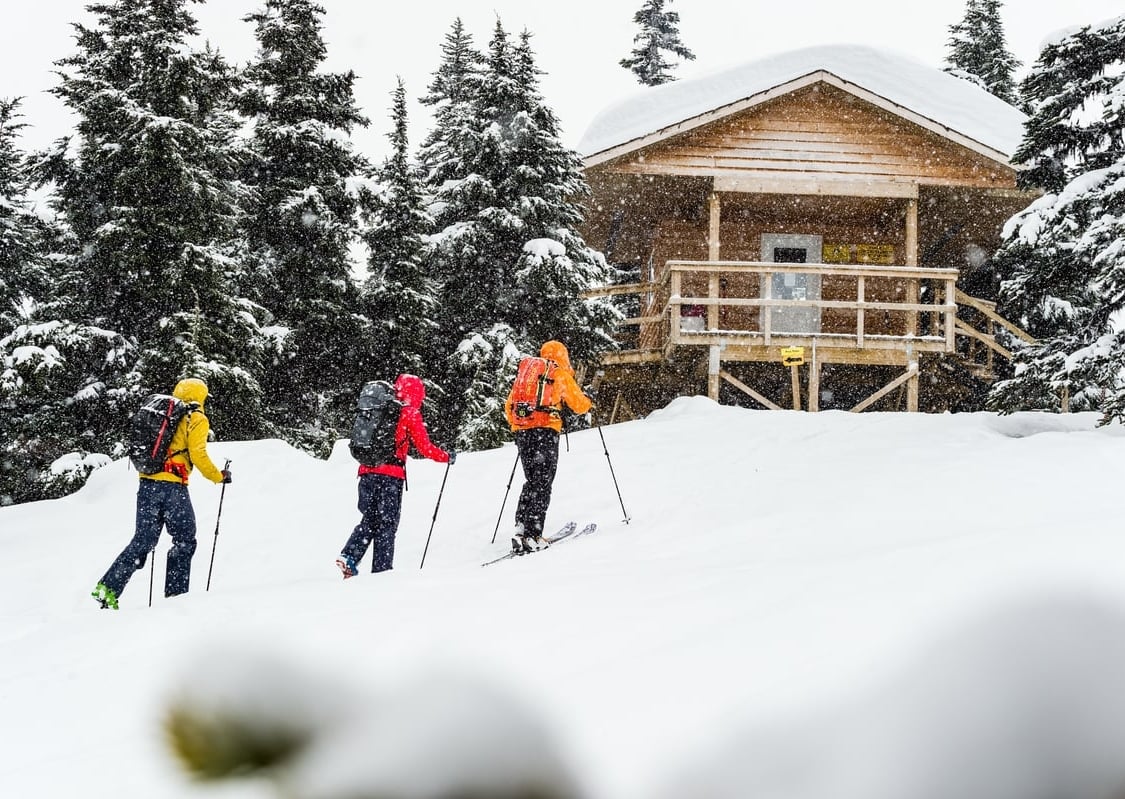 Tres esquiadores suben con pieles hasta una cabaña en la nieve rodeada de árboles. Uno lleva una chamarra amarilla, otro roja y otro naranja. Cada uno lleva una mochila.