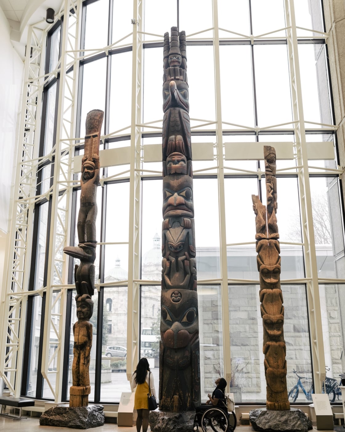 Two people at the Royal BC Museum, one standing, one in a wheelchair, gaze up at a collection of totem polls in front of a large bay window.