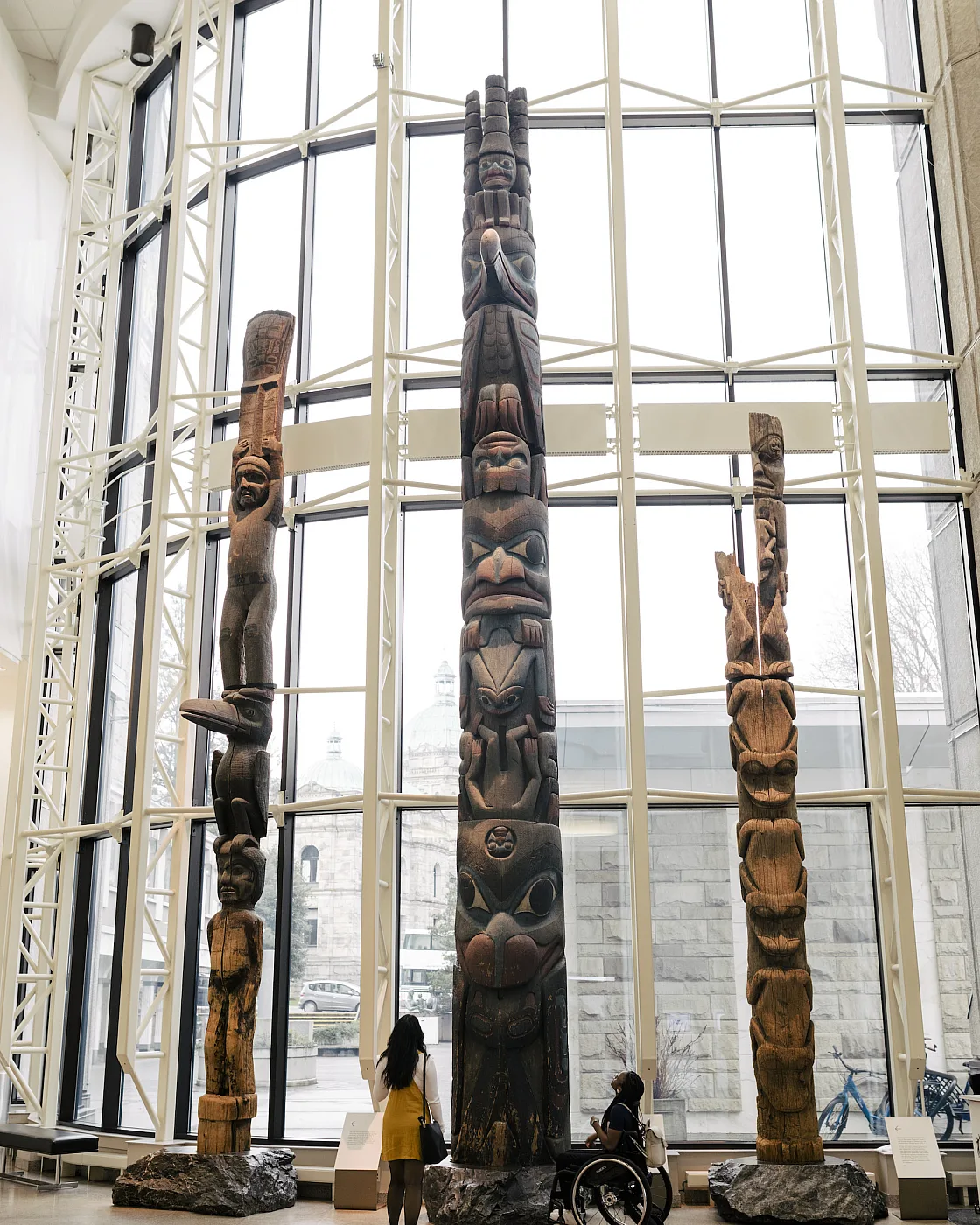 Zwei Personen im Royal BC Museum, eine stehend, eine im Rollstuhl, blicken auf eine Sammlung von Totempfählen vor einem großen Erkerfenster.