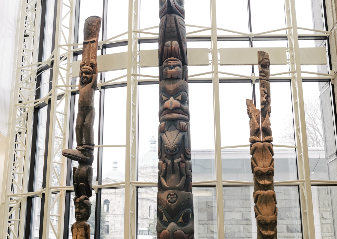 Dos personas en el Royal BC Museum, una de pie y otra en silla de ruedas, miran una colección de tótems frente a un gran ventanal.